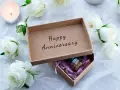 Otwarta szkatułka z napisem "Happy Anniversary" i małym prezentem w środku, otoczona białymi różami i płonącą świecą. Idealna na rocznice ślubu.
