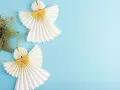 Dwa urocze aniołki diy z papieru, ozdobione złotymi gwiazdkami, wiszą na błękitnym tle obok błyszczącego, złotego listka.
