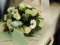 Białe róże i gerbery z zieloną wstążką na wieńcu. Na szarfie pogrzebowej widnieje napis "Kochanej Mamie".
