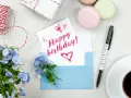 Szybkie życzenia urodzinowe: kartka z napisem "Happy birthday!", niebieska koperta, kwiaty, kawa i prezent.