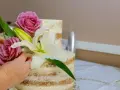 Dwie ręce dekorują dwupoziomowy tort żywymi margaretkami, różami i lilią.