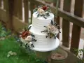 Elegancki tort weselny z białym kremem, ozdobiony bukietami kwiatów w odcieniach brązu i kremu. Wygląda jak tort z margaretkami, idealny na rustykalne przyjęcie.