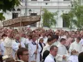 Procesja religijna z monstrancją, tłum ludzi w białych szatach, budynek w tle.