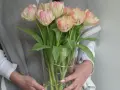 Delikatne, kremowo-różowe kwiaty podobne do tulipanów w szklanym wazonie, trzymane przez osobę w szarym swetrze.