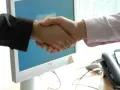 Uścisk dłoni przed monitorem komputera. Dowiedz się, jak napisać podziękowanie po udanej transakcji.