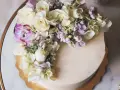 Elegancki tort z kremową polewą, ozdobiony bukietem żywych goździków, róż i hortensji w pastelowych kolorach.