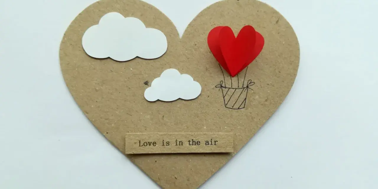 Serce z balonów w kształcie serca, unoszące się wśród chmur. Napis "Love is in the air".