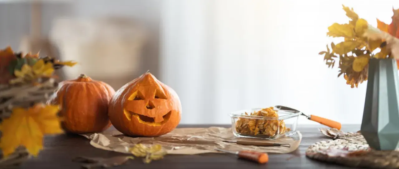 Wesoła dynia z wyciętą buzią i jej koleżanka czekają na Halloween. W szklanym naczyniu pestki dyni, gotowe na nowe pomysły na dynie.
