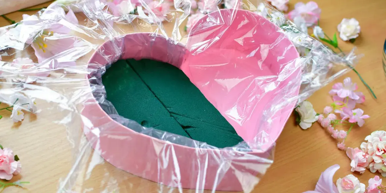 Jak zrobić flower box z winem? Różowe serce wyłożone folią i gąbką florystyczną, otoczone sztucznymi kwiatami.