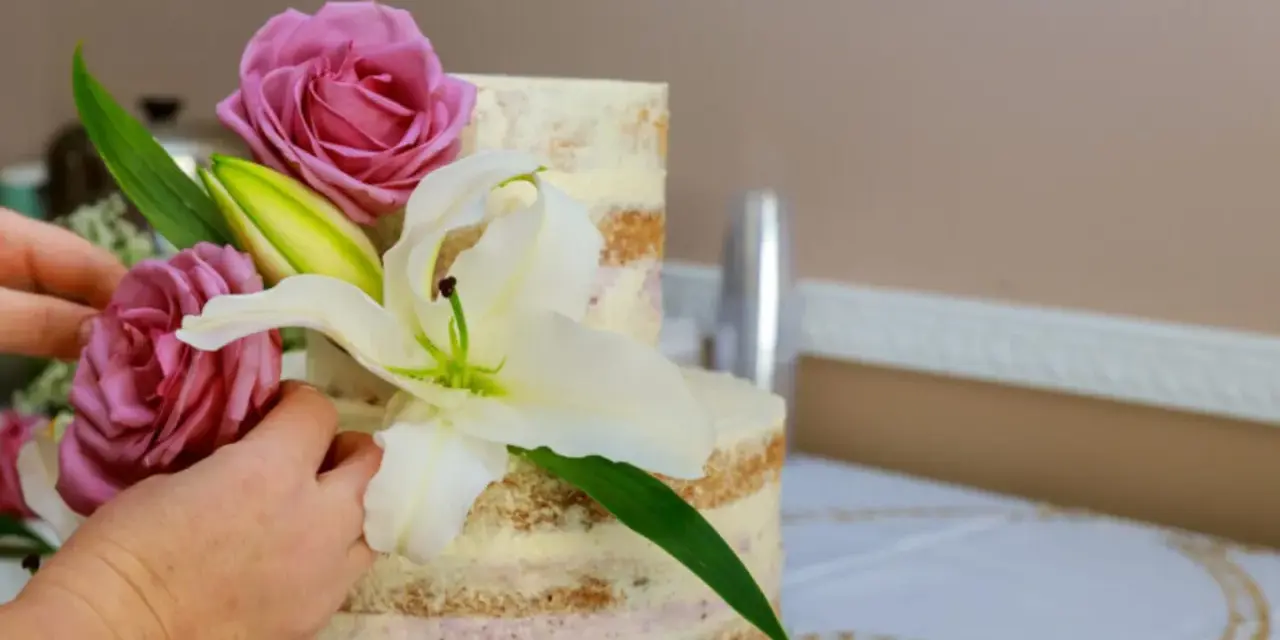 Dwie ręce dekorują dwupoziomowy tort żywymi margaretkami, różami i lilią.
