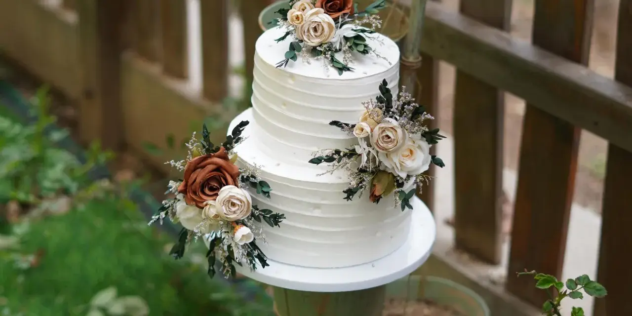 Elegancki tort weselny z białym kremem, ozdobiony bukietami kwiatów w odcieniach brązu i kremu. Wygląda jak tort z margaretkami, idealny na rustykalne przyjęcie.