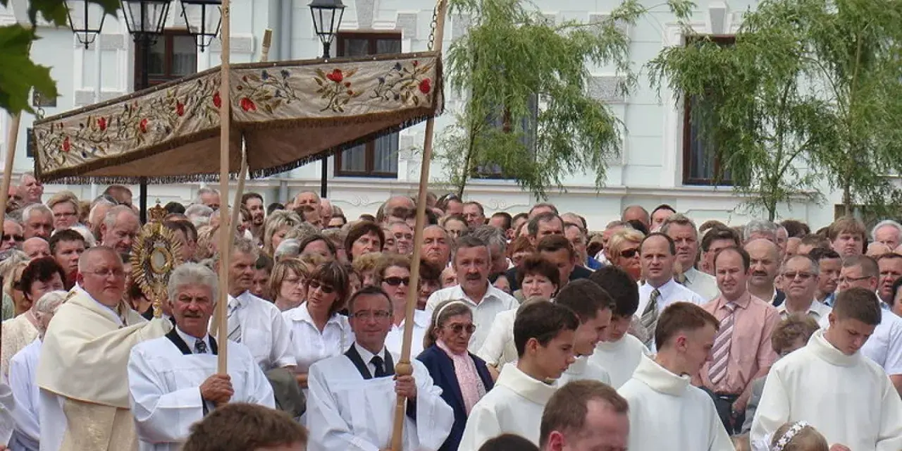 Procesja religijna z monstrancją, tłum ludzi w białych szatach, budynek w tle.