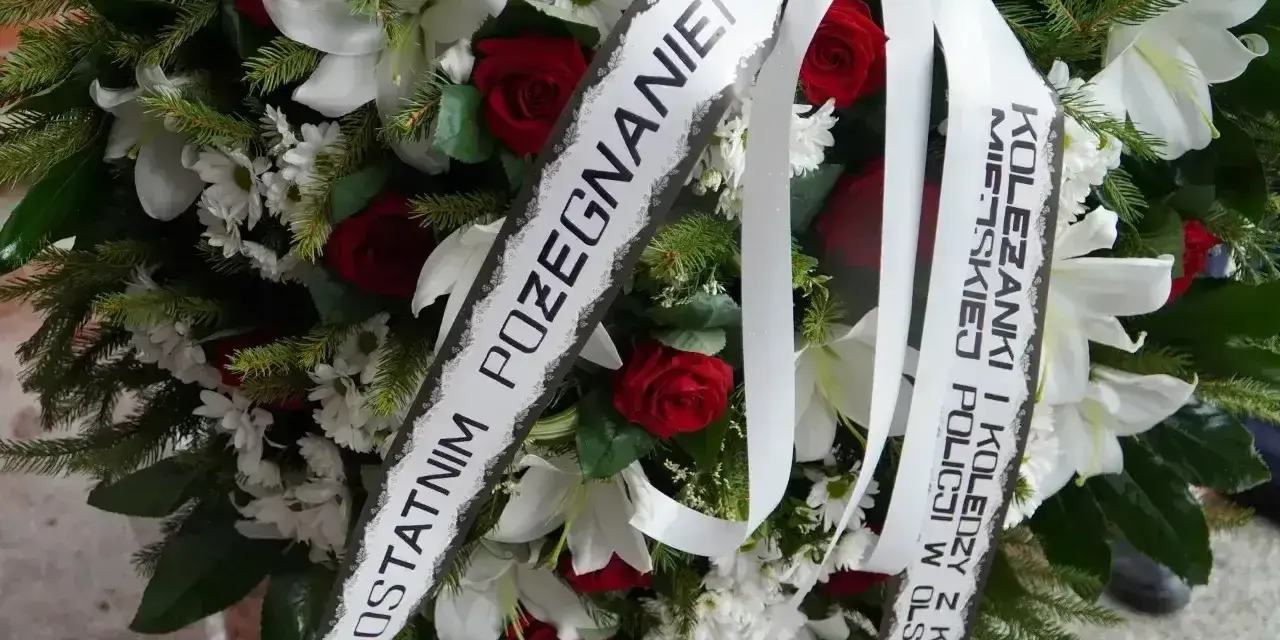 Wieniec pogrzebowy z białymi liliami i czerwonymi różami. Napis na szarfie: "Z ostatnim pożegnaniem od koleżanki i kolegów z Komendy Miejskiej Policji w Olsztynie".