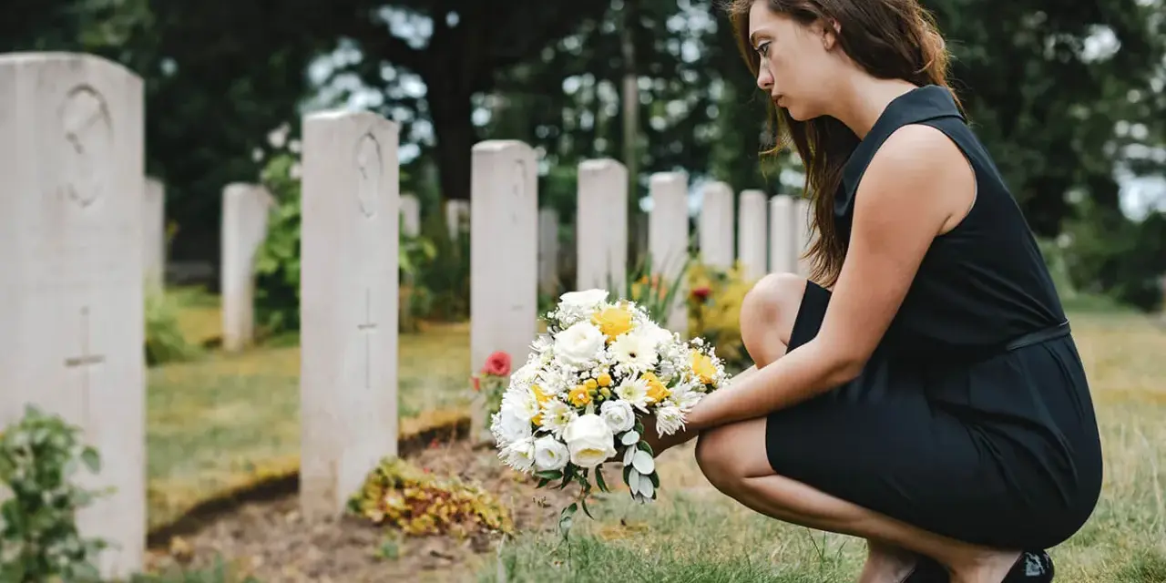 Smutna kobieta w czarnej sukience klęczy przy grobie z bukietem kwiatów. W jej myślach pewnie pojawiają się cytaty dla zmarłego taty.