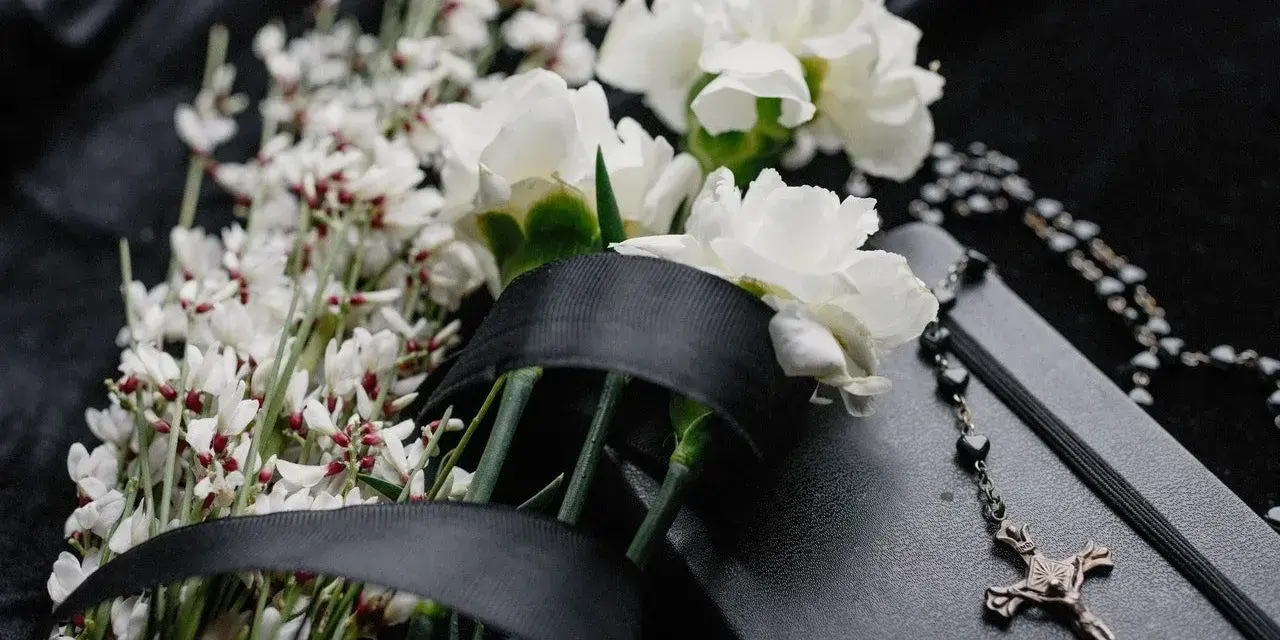 Białe kwiaty, czarna wstęga i różaniec na czarnej tkaninie. Symboliczne kwiaty na pogrzebie kolegi.