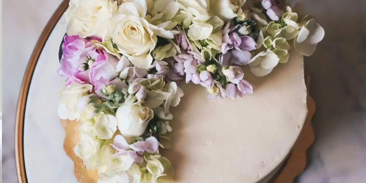 Elegancki tort z kremową polewą, ozdobiony bukietem żywych goździków, róż i hortensji w pastelowych kolorach.