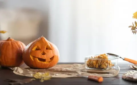 Wesoła dynia z wyciętą buzią i jej koleżanka czekają na Halloween. W szklanym naczyniu pestki dyni, gotowe na nowe pomysły na dynie.