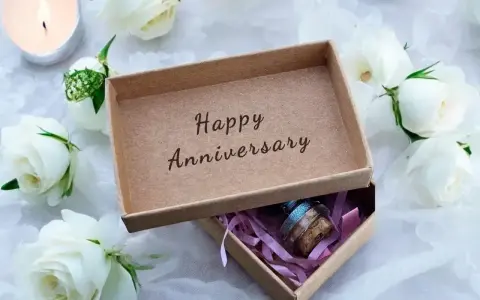 Otwarta szkatułka z napisem "Happy Anniversary" i małym prezentem w środku, otoczona białymi różami i płonącą świecą. Idealna na rocznice ślubu.