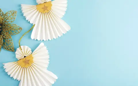 Dwa urocze aniołki diy z papieru, ozdobione złotymi gwiazdkami, wiszą na błękitnym tle obok błyszczącego, złotego listka.