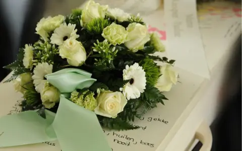 Białe róże i gerbery z zieloną wstążką na wieńcu. Na szarfie pogrzebowej widnieje napis "Kochanej Mamie".