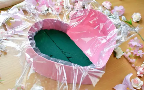 Jak zrobić flower box z winem? Różowe serce wyłożone folią i gąbką florystyczną, otoczone sztucznymi kwiatami.