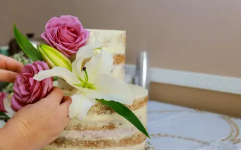Dwie ręce dekorują dwupoziomowy tort żywymi margaretkami, różami i lilią.