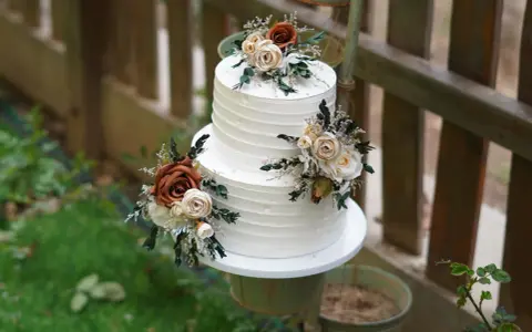 Elegancki tort weselny z białym kremem, ozdobiony bukietami kwiatów w odcieniach brązu i kremu. Wygląda jak tort z margaretkami, idealny na rustykalne przyjęcie.