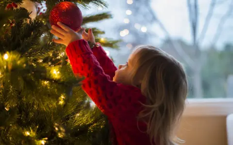 Mała dziewczynka w czerwonym sweterku wiesza czerwoną bombkę na choince. To jej imieniny 24.12, czas świątecznych przygotowań.