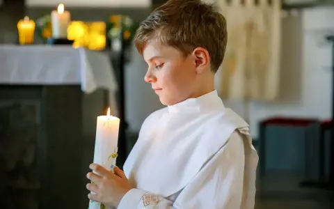 Chłopiec w bieli trzyma zapaloną świecę podczas uroczystości chrztu świętego.