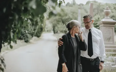 Smutek i wsparcie na cmentarzu. Kobieta ociera łzy, mężczyzna obejmuje ją ramieniem. Ich obecność na pogrzebie jest świadectwem głębokiej więzi.