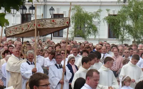 Procesja religijna z monstrancją, tłum ludzi w białych szatach, budynek w tle.