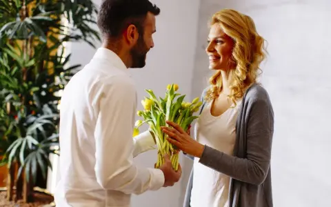 Mężczyzna wręcza kobiecie bukiet żółtych kwiatów. Uśmiechnięta kobieta z blond włosami przyjmuje prezent.