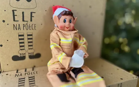 Uroczy elf na półce w stroju strażaka siedzi na kartonie z rysunkami elfich nóg.