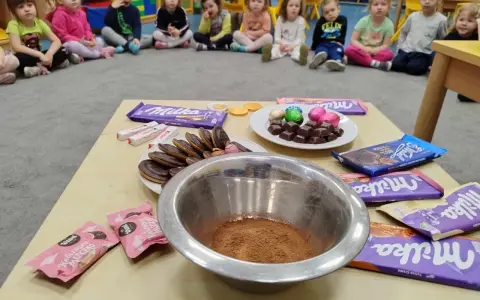 Dzieci czekają na słodkie pomysły na dzień czekolady. Na stole czekają czekolady Milka, Kinder i ciastka.