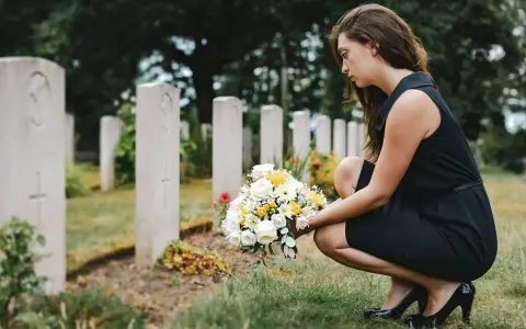 Smutna kobieta w czarnej sukience klęczy przy grobie z bukietem kwiatów. W jej myślach pewnie pojawiają się cytaty dla zmarłego taty.