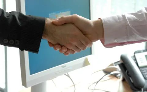 Uścisk dłoni przed monitorem komputera. Dowiedz się, jak napisać podziękowanie po udanej transakcji.