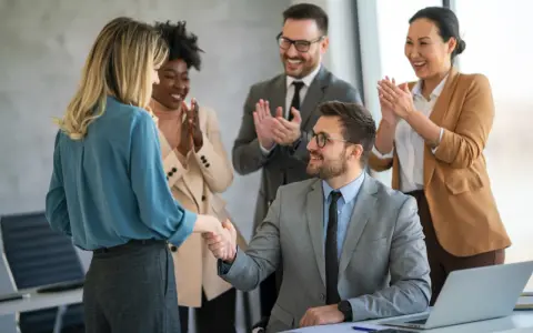 Zespół gratuluje pracownicy, ściskając jej dłoń. Wszyscy uśmiechnięci, to wyraz podziękowania za zaangażowanie.