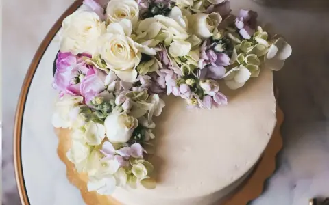 Elegancki tort z kremową polewą, ozdobiony bukietem żywych goździków, róż i hortensji w pastelowych kolorach.