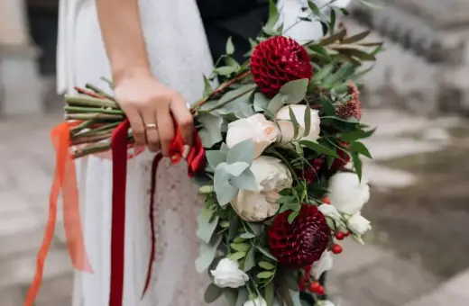 Panna kwiatowa trzyma bukiet z czerwonych dalii, białych róż i zielonych liści, ozdobiony wstążkami.