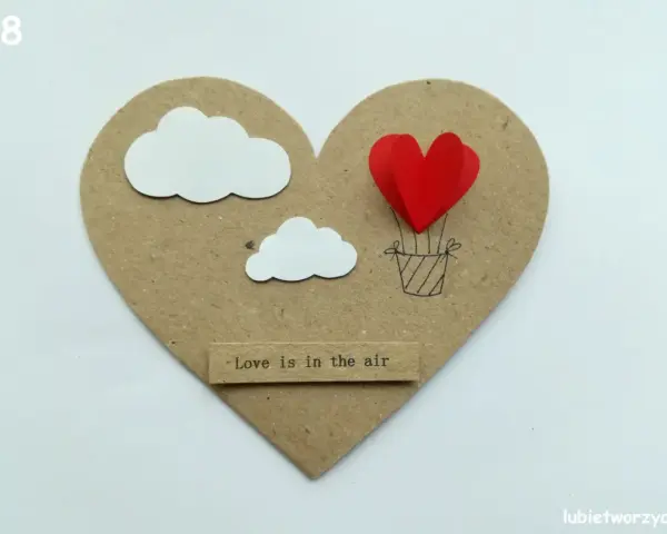 Serce z balonów w kształcie serca, unoszące się wśród chmur. Napis "Love is in the air".