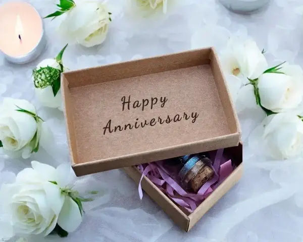 Otwarta szkatułka z napisem "Happy Anniversary" i małym prezentem w środku, otoczona białymi różami i płonącą świecą. Idealna na rocznice ślubu.