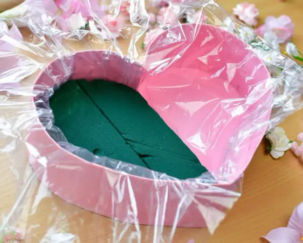 Jak zrobić flower box z winem? Różowe serce wyłożone folią i gąbką florystyczną, otoczone sztucznymi kwiatami.