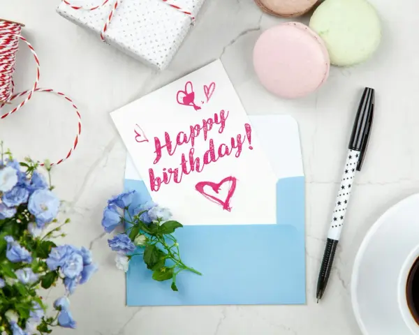 Szybkie życzenia urodzinowe: kartka z napisem "Happy birthday!", niebieska koperta, kwiaty, kawa i prezent.