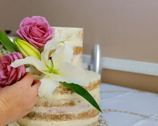 Dwie ręce dekorują dwupoziomowy tort żywymi margaretkami, różami i lilią.