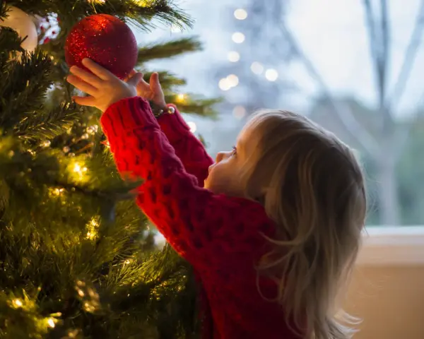 Mała dziewczynka w czerwonym sweterku wiesza czerwoną bombkę na choince. To jej imieniny 24.12, czas świątecznych przygotowań.