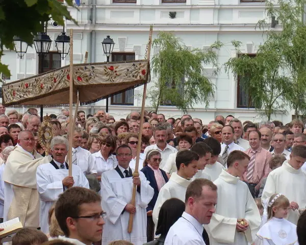 Procesja religijna z monstrancją, tłum ludzi w białych szatach, budynek w tle.