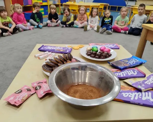 Dzieci czekają na słodkie pomysły na dzień czekolady. Na stole czekają czekolady Milka, Kinder i ciastka.