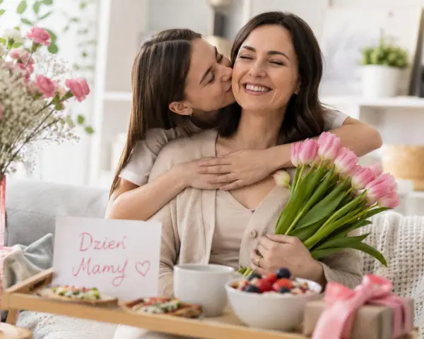 Córka całuje mamę w policzek, wręczając jej bukiet różowych tulipanów i kartkę "Dzień Mamy". 26.05 imieniny.