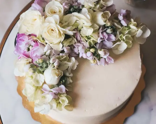 Elegancki tort z kremową polewą, ozdobiony bukietem żywych goździków, róż i hortensji w pastelowych kolorach.