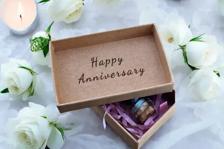 Otwarta szkatułka z napisem "Happy Anniversary" i małym prezentem w środku, otoczona białymi różami i płonącą świecą. Idealna na rocznice ślubu.